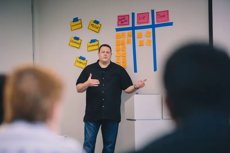 A Mountain Goat Software trainer leading a class beside a Scrum values and task board